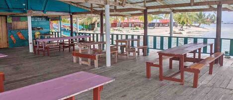 Un área de comedor al aire libre con mesas y bancos de madera, techo cubierto y vista a la playa y al océano.