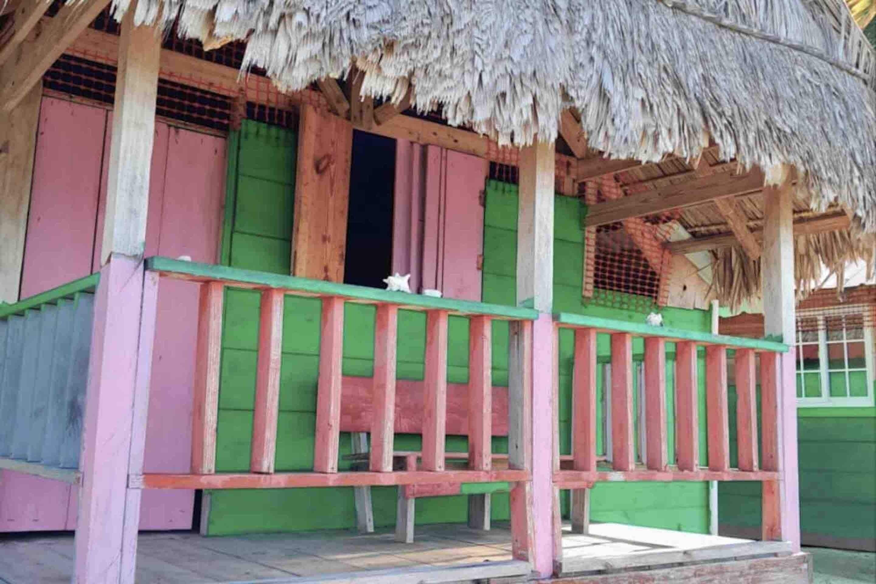 Una estructura de madera con techo de paja, paredes verdes y rosadas, y un banco de madera.