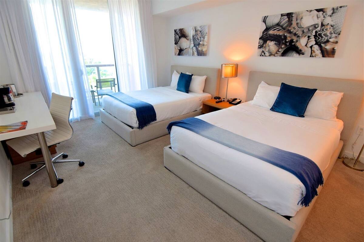 A hotel room with two beds, a desk, and a chair. There are paintings on the walls and a lamp on the nightstand.