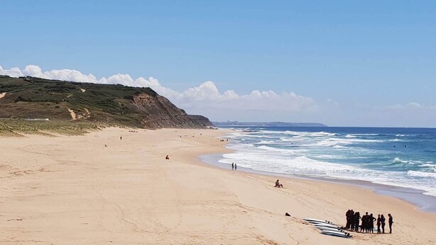 A wide sandy beach with gentle waves, a grassy hill in the background, and a clear sky. - Harmonic House with sea view (Atalaia Lourinhã)