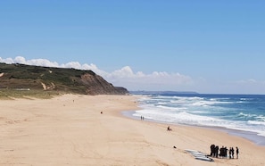 A wide sandy beach with gentle waves, a grassy hill in the background, and a clear sky.