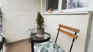 A small balcony with a round table and a wooden chair, a potted plant, and a window overlooking a tree.