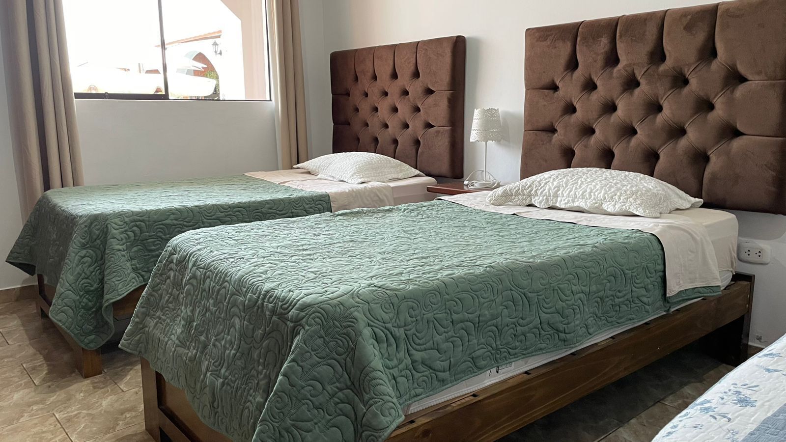 Two single beds with tufted headboards, a bedside table with a lamp, and a window with curtains.