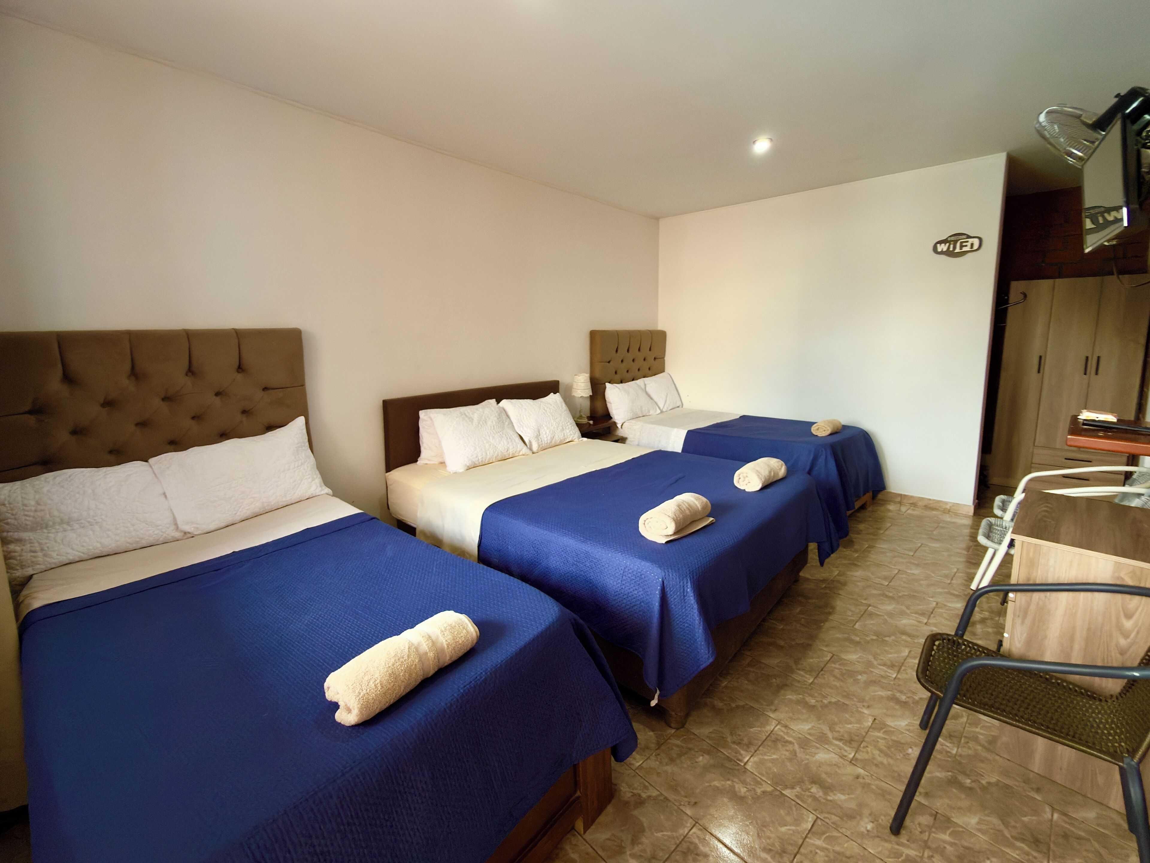 A hotel room with three beds, each with a blue bedspread and a rolled towel. There is a wooden chair and a small table with a lamp.