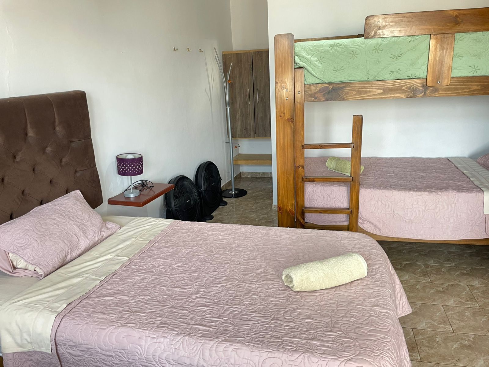 A room with a large bed, a smaller bed, a wooden ladder, a desk with a lamp, and two fans.