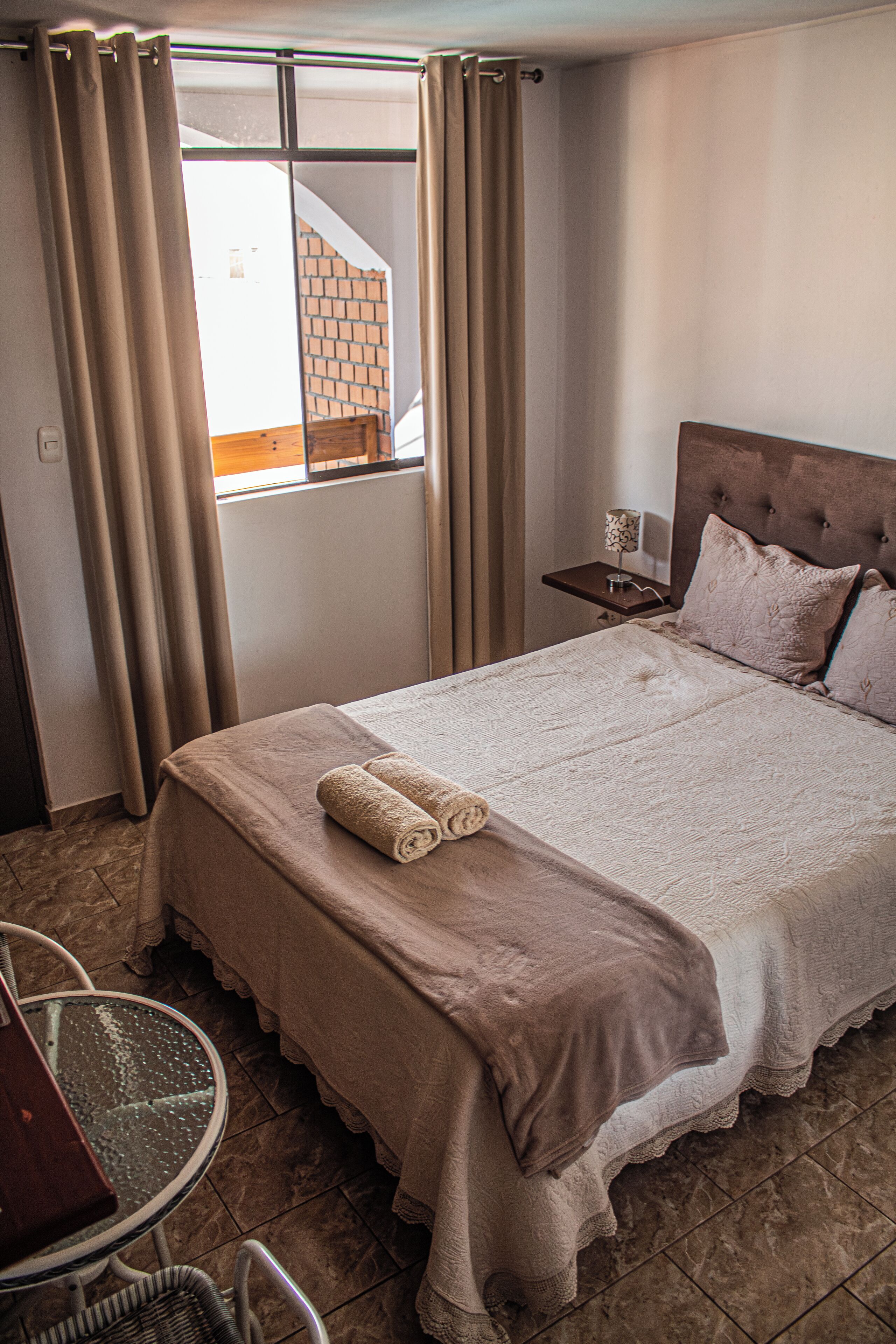 A neatly made bed with a brown bedspread and pillows, a small bedside table with a lamp, and a view of a brick building through a window with curtains.