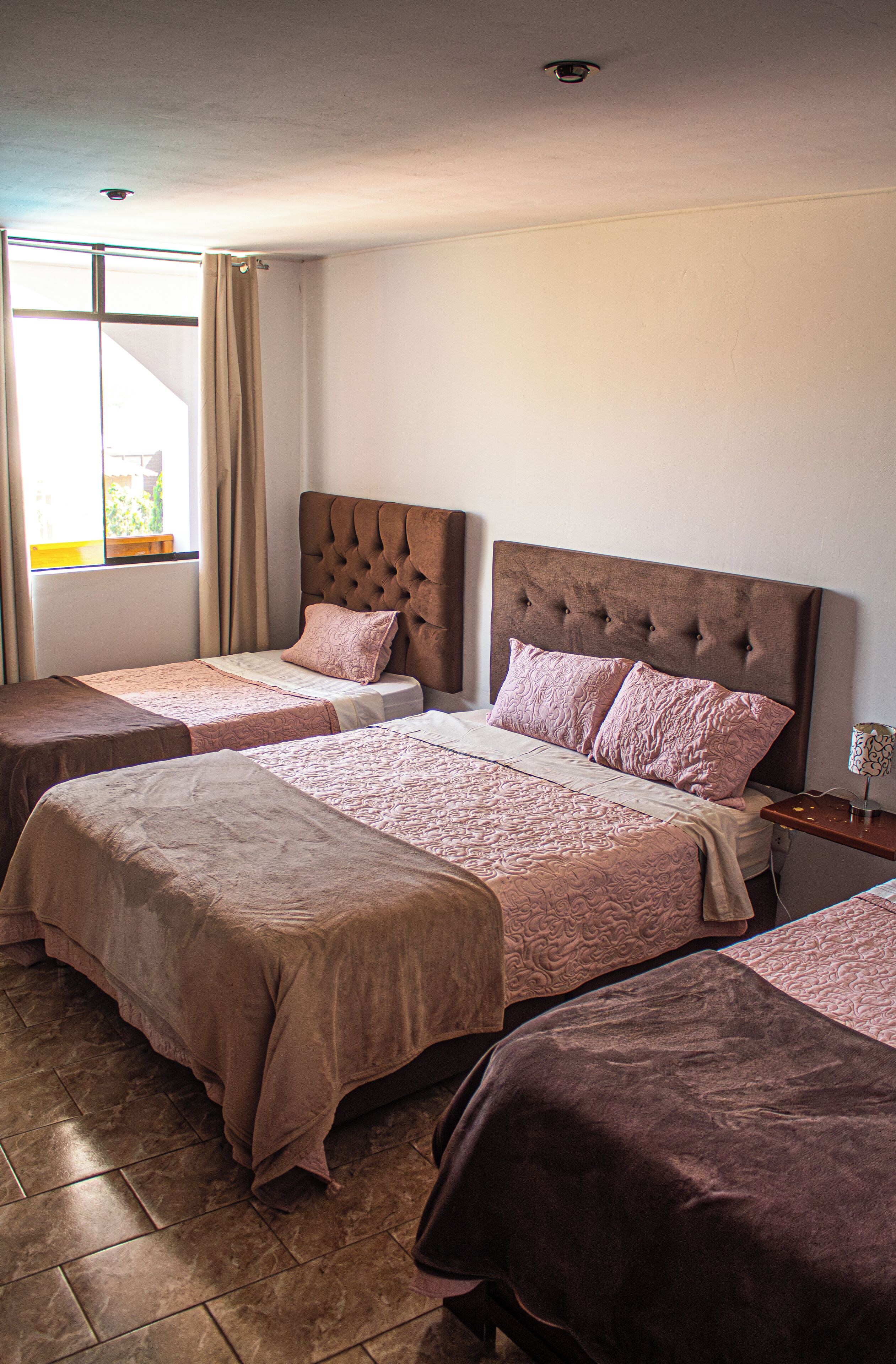 Habitación de hotel con dos camas, una ventana con cortinas, una mesita de noche con lámpara y suelo de baldosas.