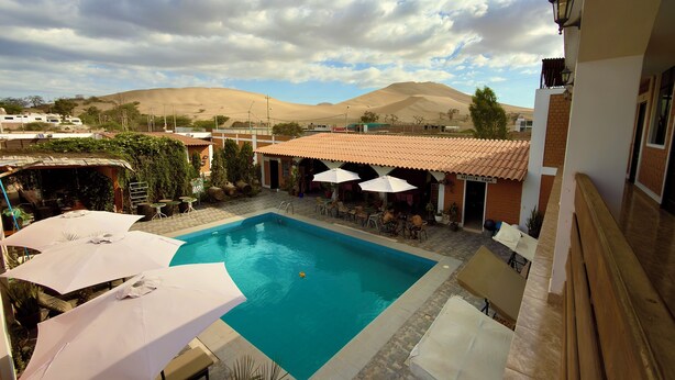 A pool area with lounge chairs and umbrellas, a building with a tiled roof, and a mountainous backdrop.