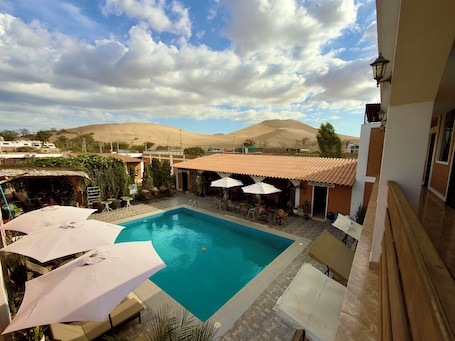 A pool area with lounge chairs and umbrellas, a building with a tiled roof, and a mountainous backdrop.