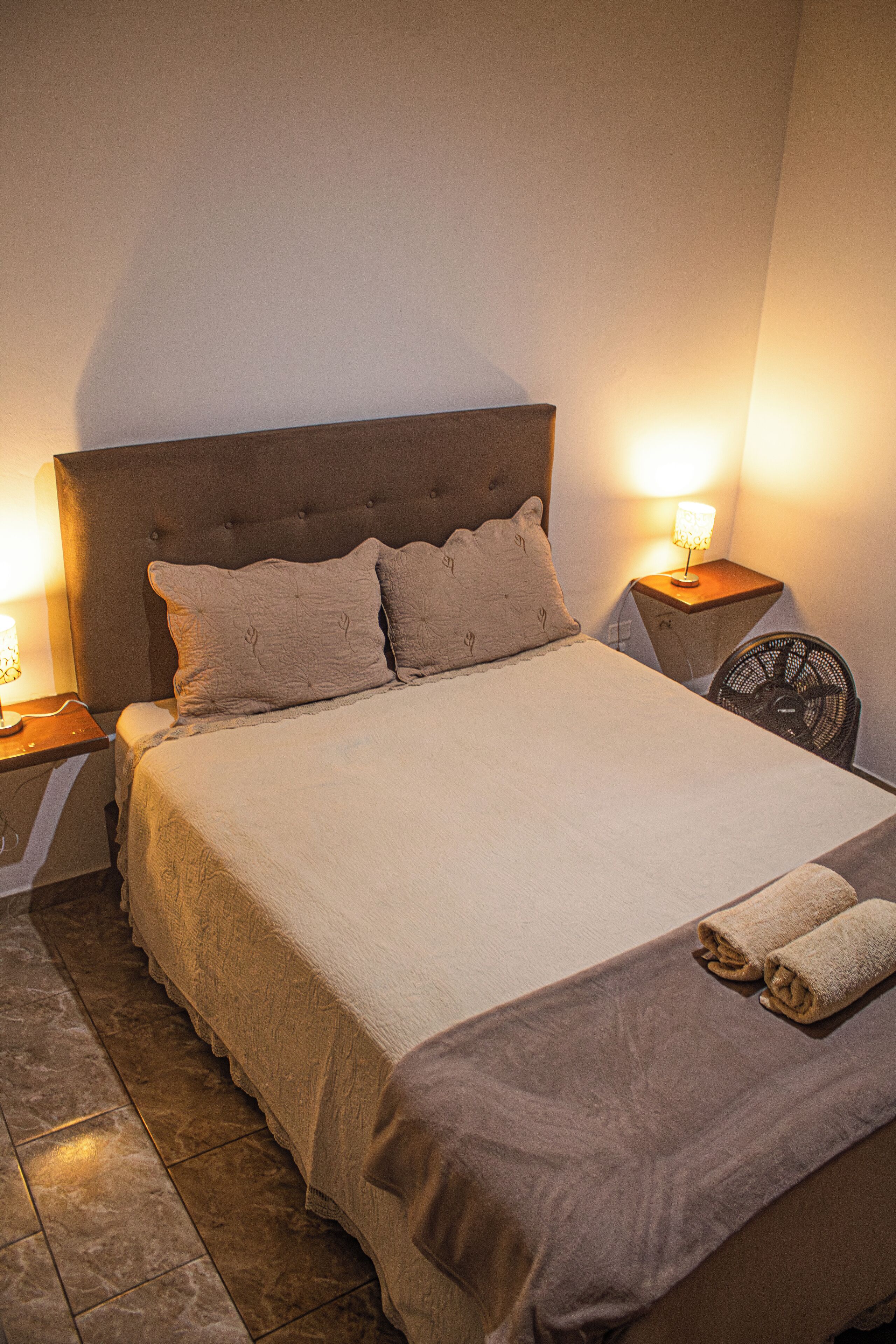 Un lit bien fait, avec une tête de lit, deux lampes de chevet et un petit ventilateur posé sur une table de nuit.