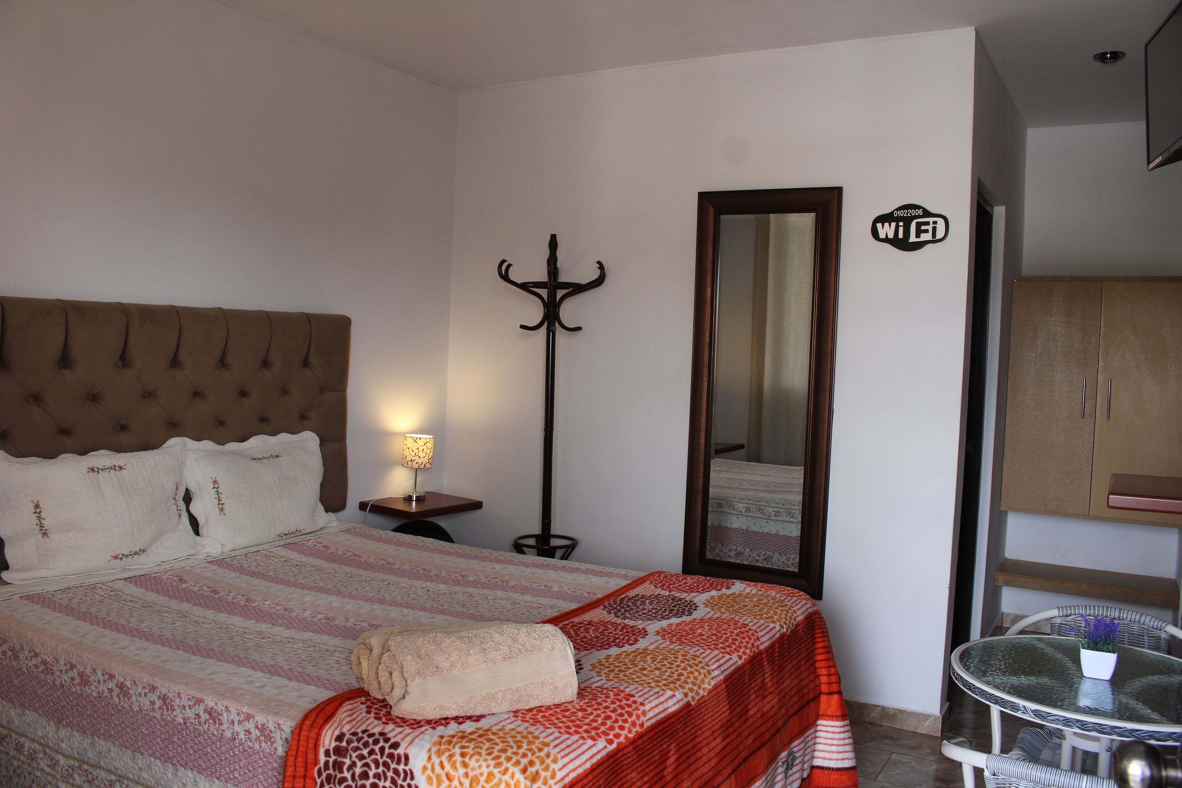 A bedroom with a bed, a bedside table with a lamp, a coat rack, a mirror, and a small table with a chair.