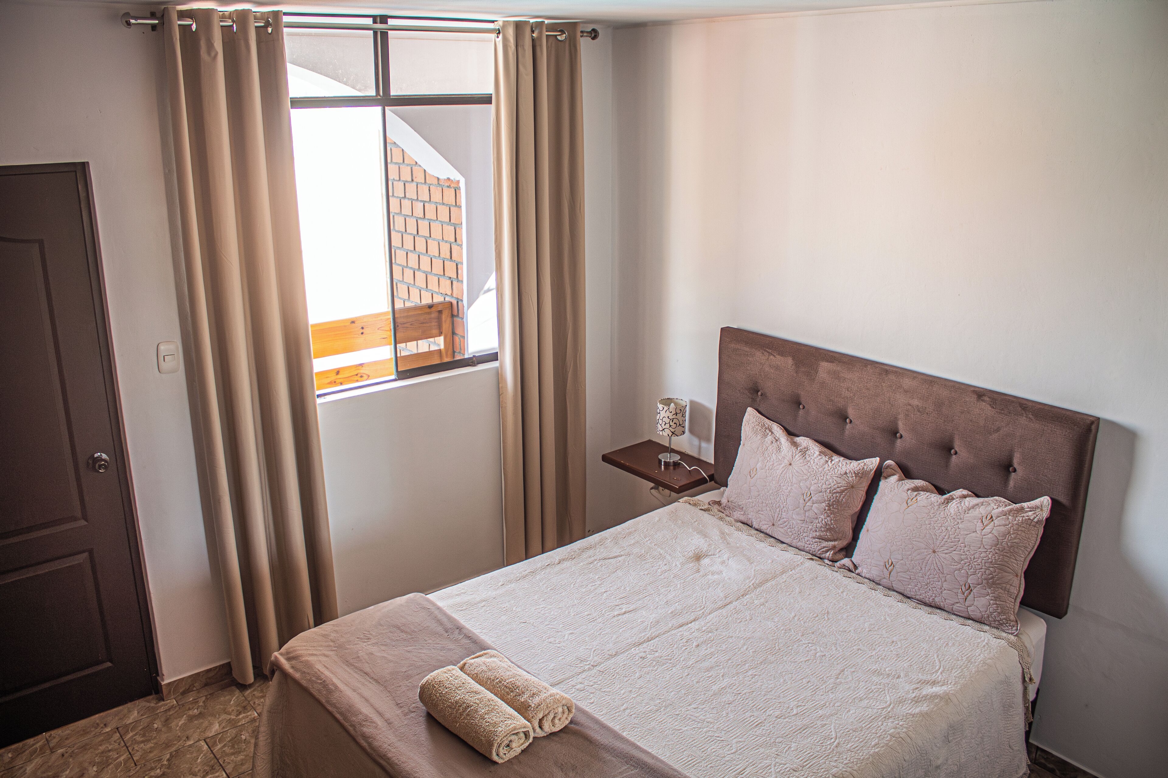 A neatly made bed with a headboard, a small bedside table with a lamp, and a window with curtains.