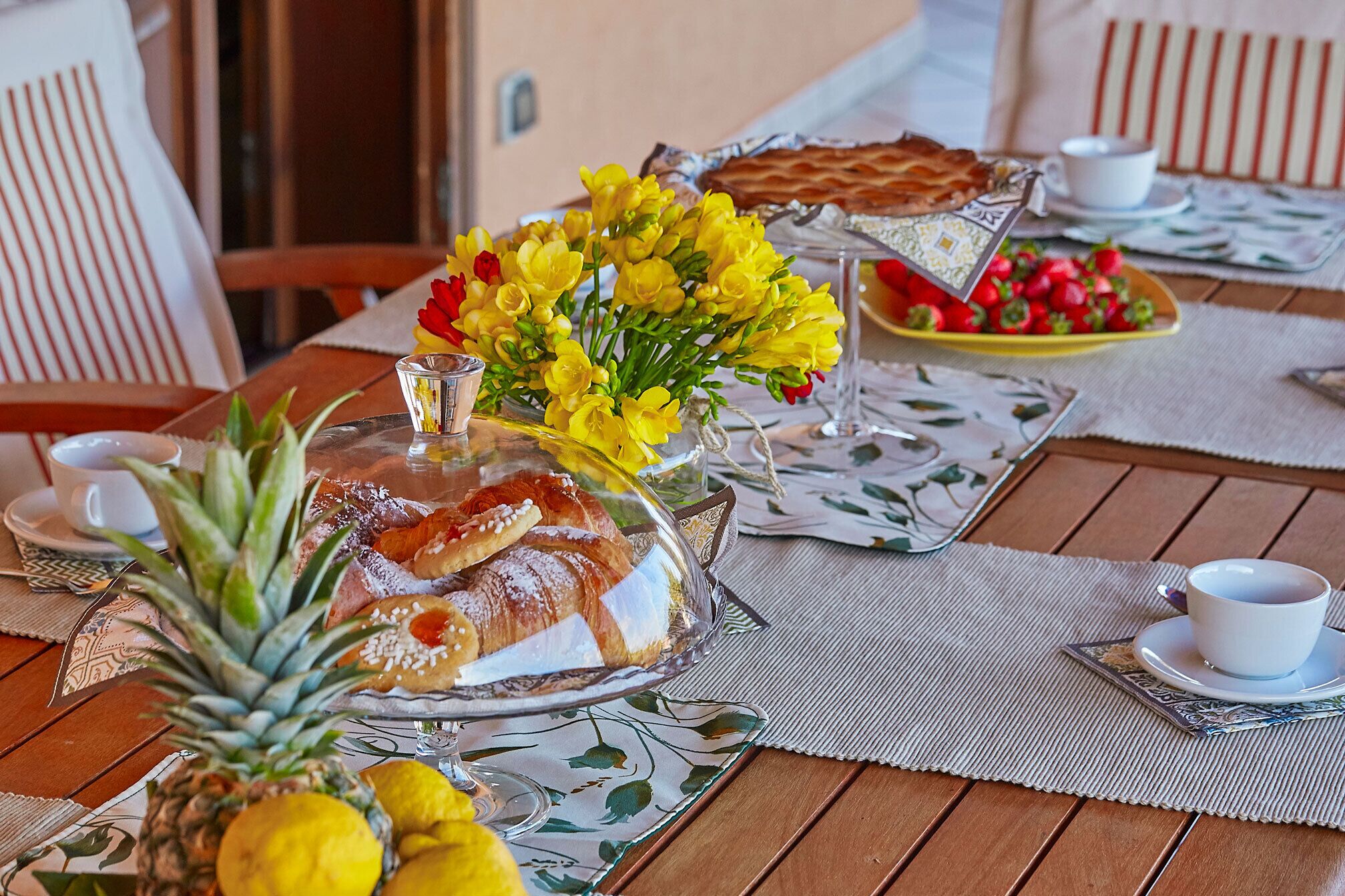 A breakfast setting with a covered cake, a vase of yellow flowers, and a plate of strawberries.