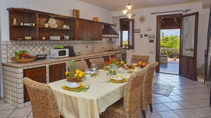 Un comedor con una mesa dispuesta para cuatro personas, rodeado de sillas de mimbre. Se ve una cocina con armarios de madera y un salpicadero de azulejos al fondo.