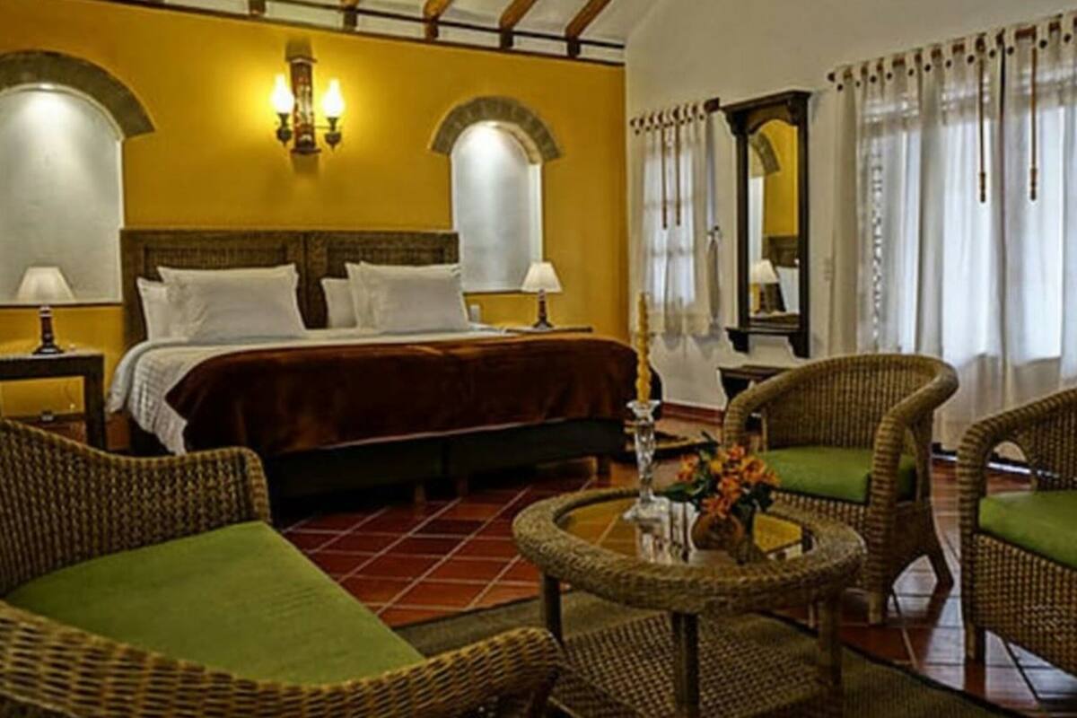 A hotel room with a bed, two nightstands, a round coffee table, wicker chairs with green cushions, and a large mirror.
