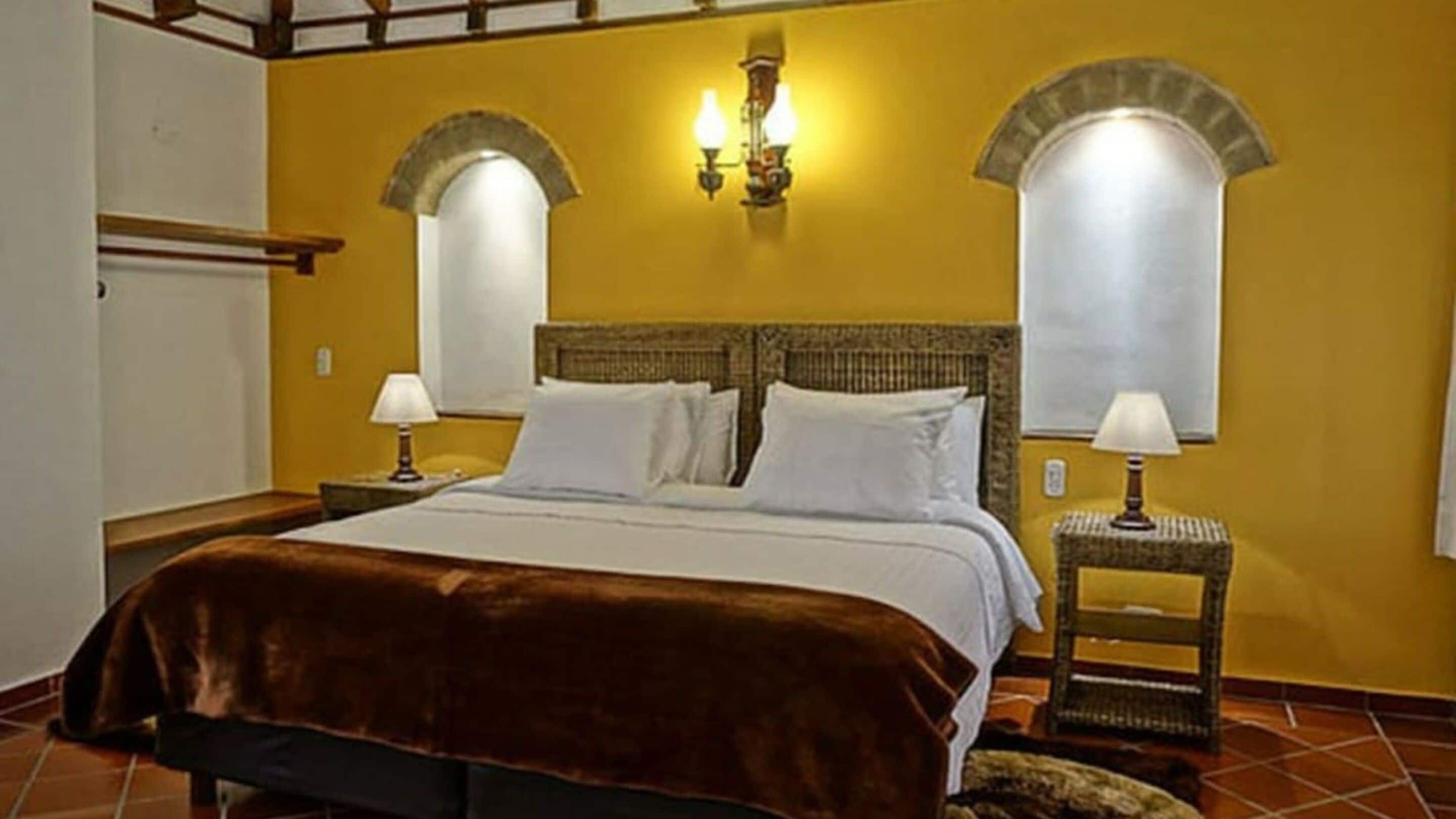 Habitación de hotel con cama, dos mesitas de noche, una lámpara de pared y una ventana con cortina.