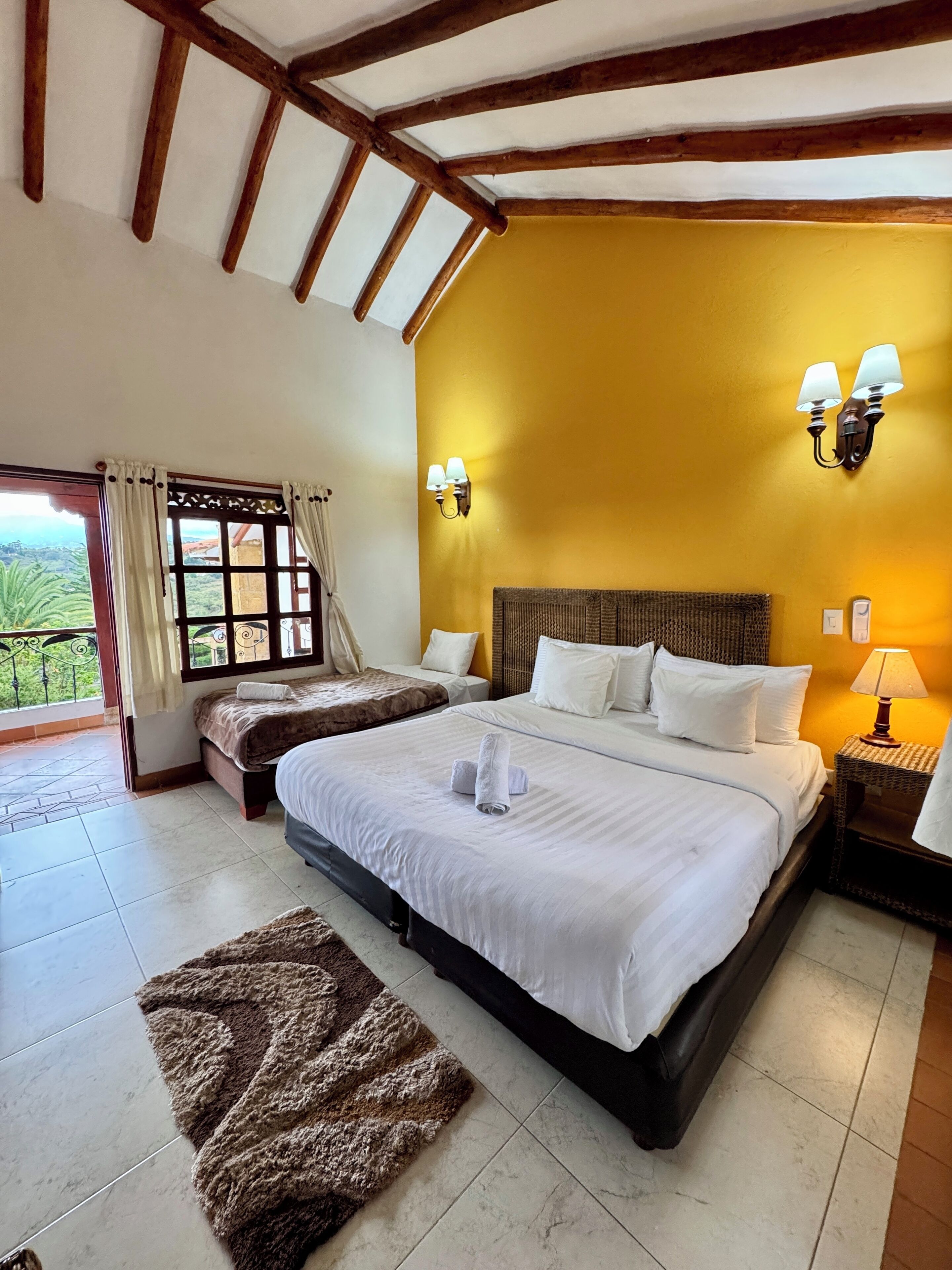 A bedroom with a large bed, two bedside tables with lamps, a window with curtains, and a view of greenery outside.