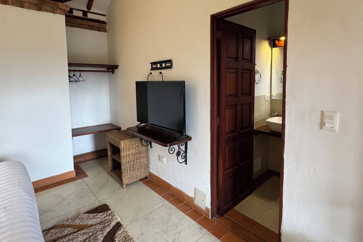A room with a bed, a TV on a stand, a wooden bench, a woven basket, and a door leading to a bathroom.