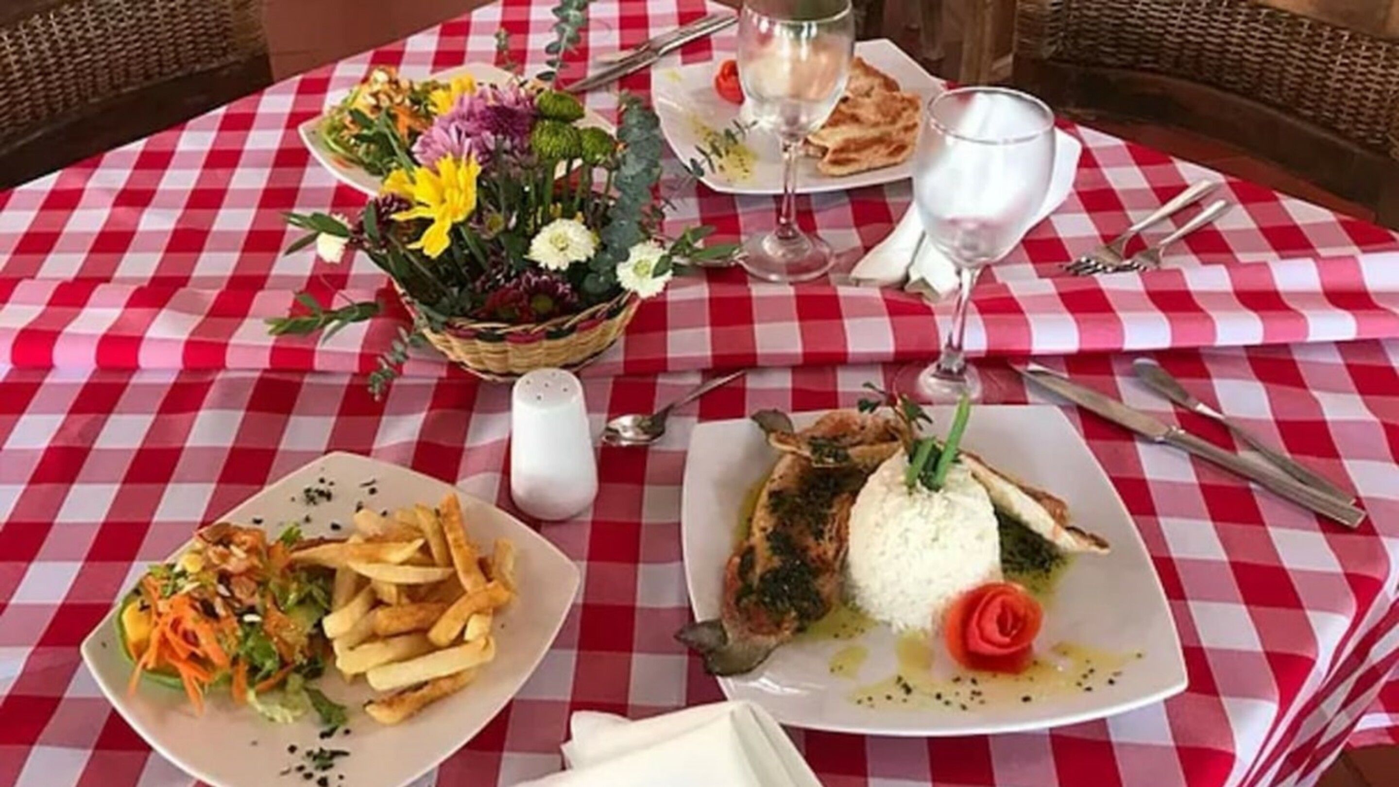 A table set with a red and white checkered tablecloth, plates of food, a basket of flowers, and cutlery.