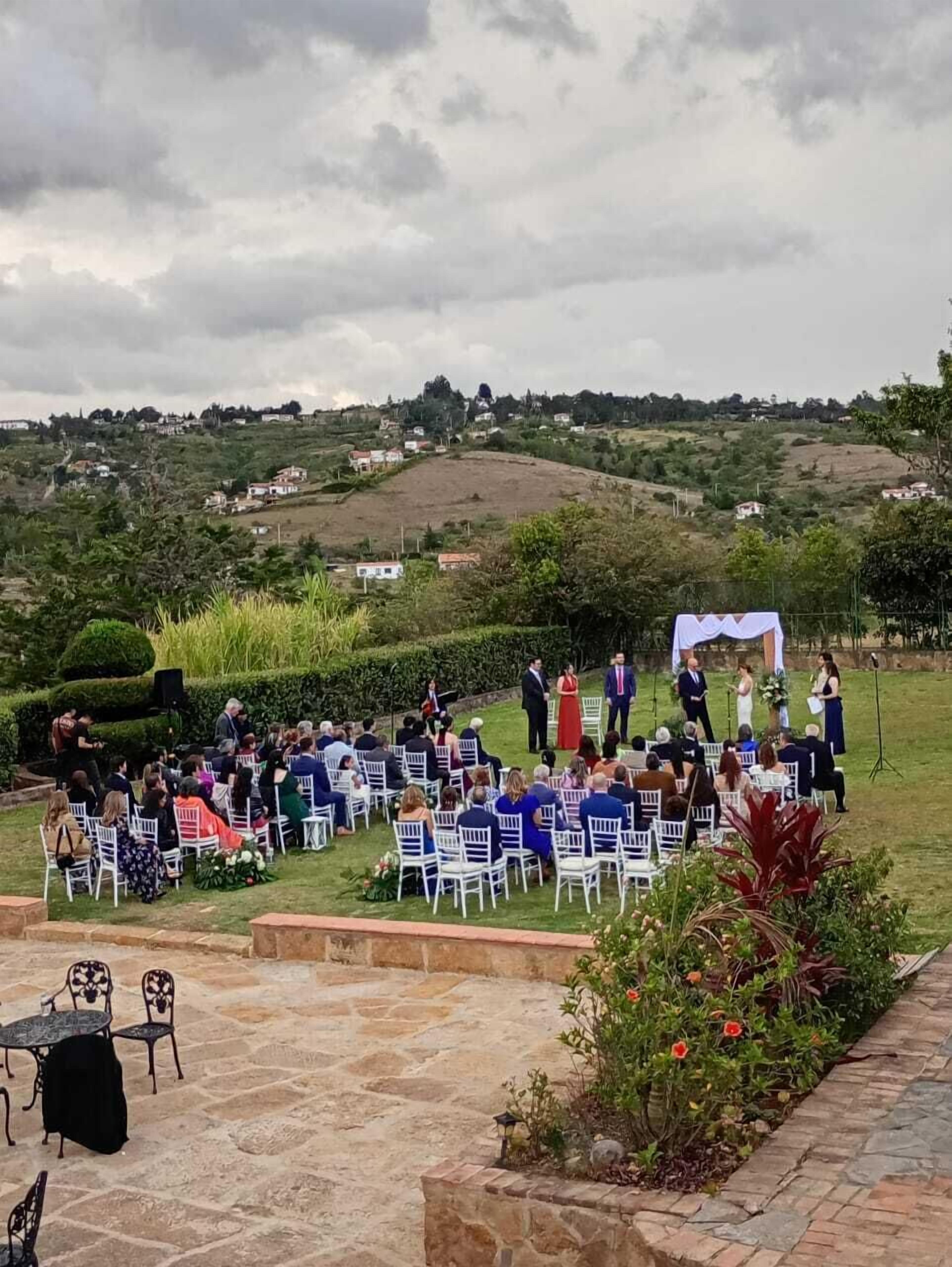 An outdoor wedding ceremony with guests seated on white chairs arranged in rows, overlooking a hilly landscape with scattered buildings.