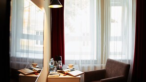 Una camera d'albergo con scrivania, sedia, letto e ampie finestre con tende leggere.