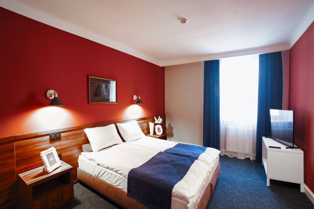 Una habitación de hotel con cabecera de madera, cama con sábanas blancas, mesita de noche con lámpara, televisor sobre un mueble bajo y ventana con cortinas azules.