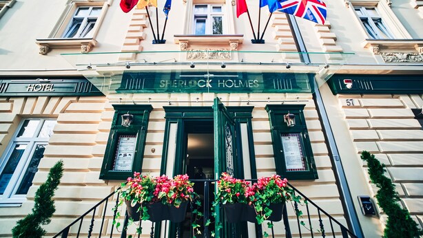 Ingresso di un hotel con bandiere, un cartello con su scritto "Sherlock Holmes" e fioriere.