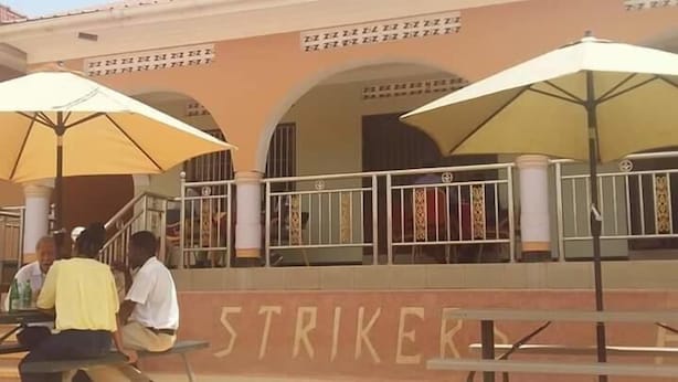 A building with the word "STRIKER" on the wall, outdoor seating with umbrellas, and a gated entrance. - Strikers Hotel (Soroti)