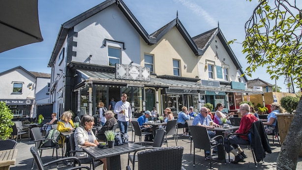 Ulkoterassi, jossa on pöytiä ja tuoleja, rakennus, jonka yläpuolella on kyltti, jossa lukee ”Café Bar N’ Bistrot”, ja taustalla näkyy kyltti, jossa lukee ”Fish Bar”.
