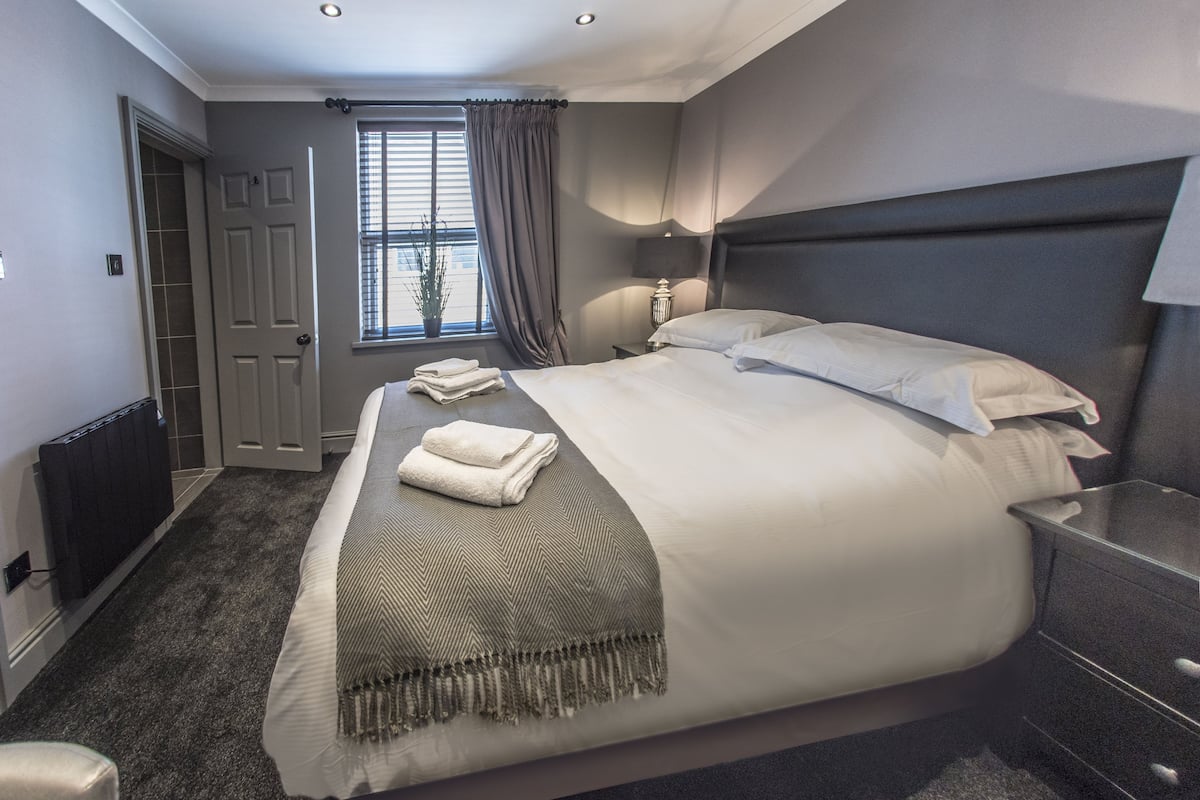 A modern bedroom with a large bed, bedside tables, a radiator, a window with curtains, and a door.