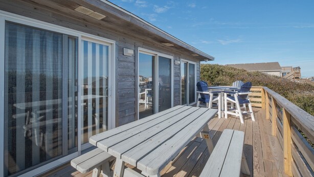 A wooden deck with a table and chairs, large sliding glass doors, and a view of the beach. - 4752- Canopy Retreat, Walk to Beach | Quiet Kitty Hawk Retreat (Kitty Hawk)