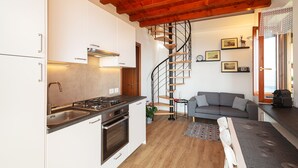 A modern kitchen with white cabinets, a built-in stove, and a spiral staircase leading to a living area with a gray sofa and a coffee table. - Panoramic View of Lake Maggiore Near Stresa • Private Garden & Parking (Levo di Stresa)