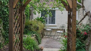 Une tonnelle en bois mène à un patio agrémenté d’une table et de chaises, le tout entouré de verdure et bordé d’un mur blanc.