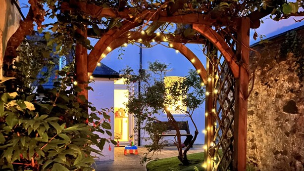Un chemin menant à un bâtiment doté d’un mur en pierre et d’une arche en bois ornée de guirlandes lumineuses.