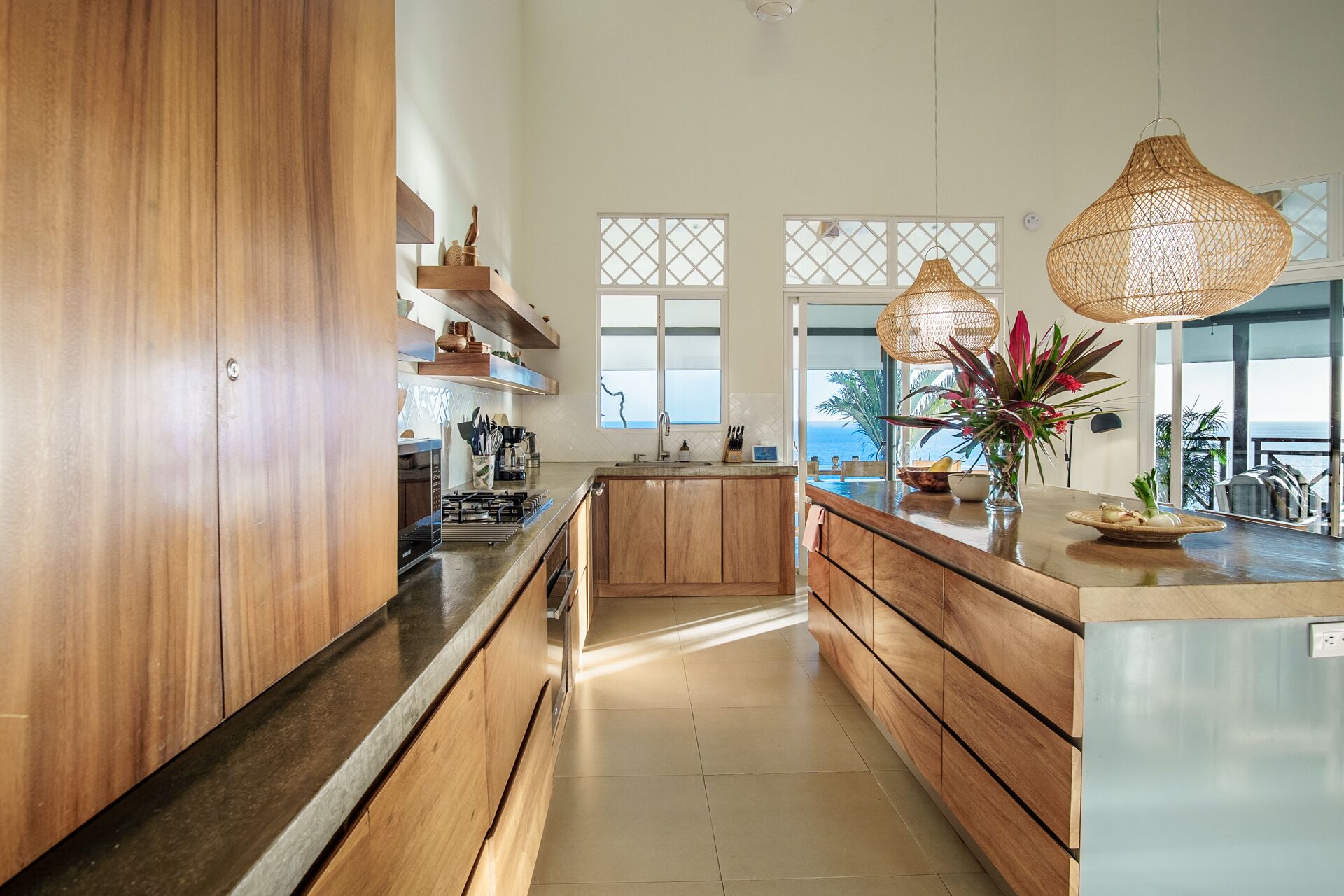 A modern kitchen with wooden cabinets, a central island, and large windows offering a view of the sea.