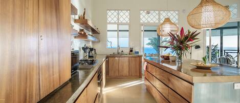 A modern kitchen with wooden cabinets, a central island, and large windows offering a view of the sea.