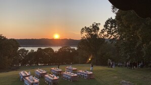 Een terras met tafels en stoelen, gelegen op een grasveld met uitzicht op een wateroppervlak tijdens de zonsondergang.