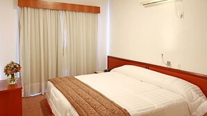 Una habitación de hotel con cama, una mesita de noche con un florero y una ventana con cortinas, además de un aire acondicionado instalado en la pared.