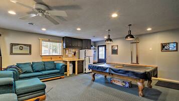 A room with a pool table, a sofa, a ceiling fan, and a kitchen area.