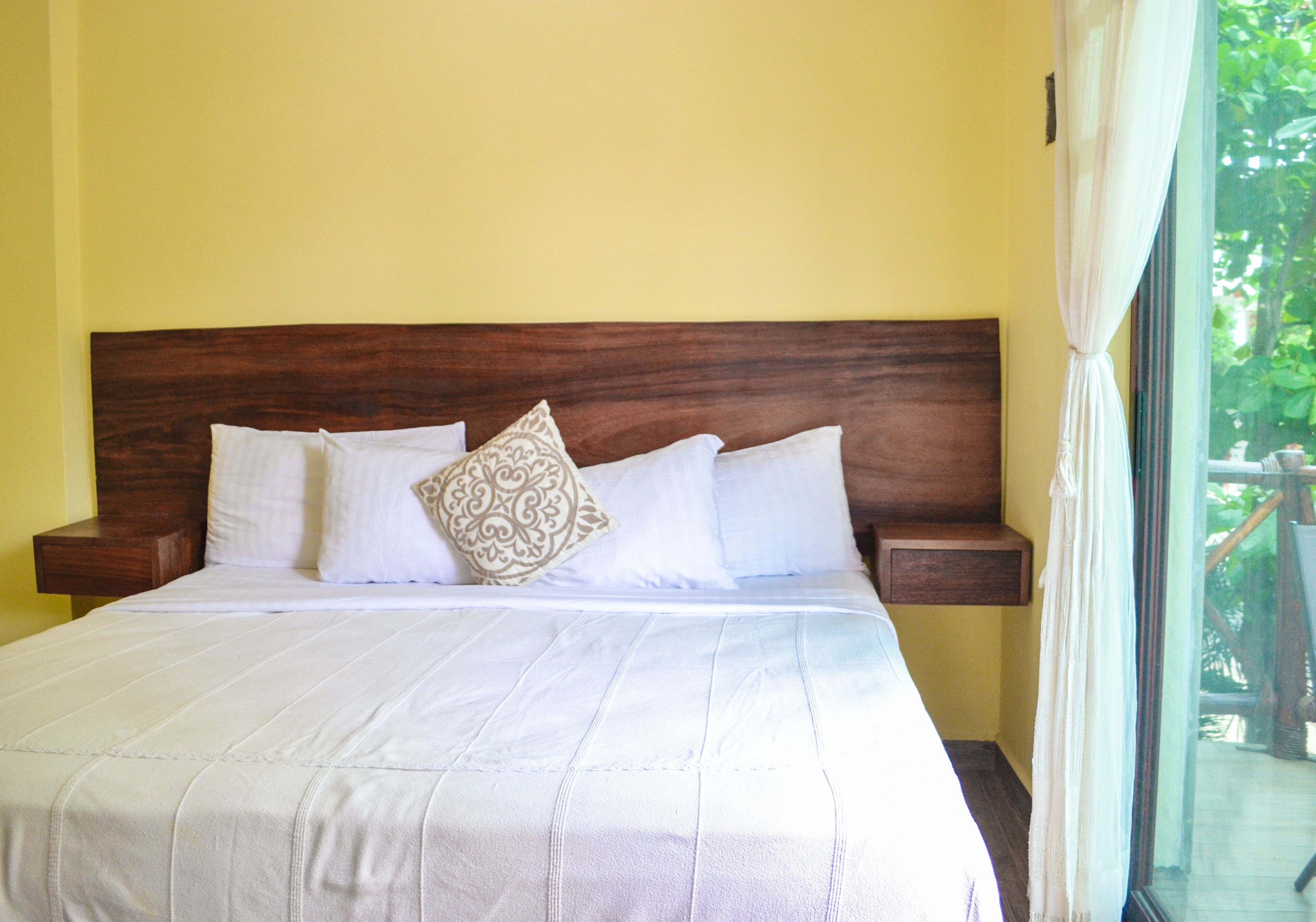 Una cama bien hecha con sábanas blancas y un cojín estampado, cabecera de madera y mesitas de noche frente a una pared amarilla.