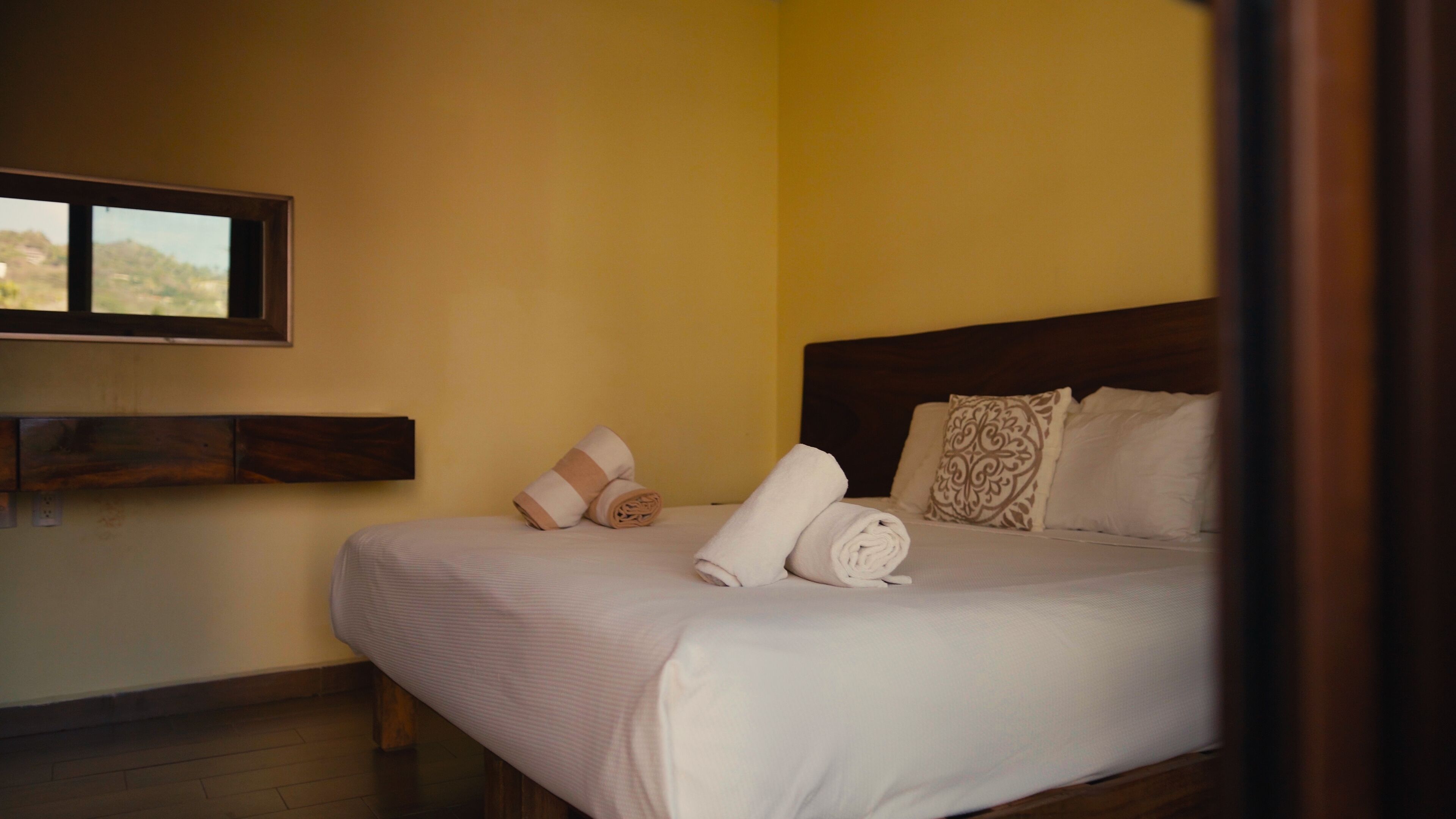 A neatly made bed with pillows and a rolled towel, a wooden headboard, a wall-mounted TV, and a window with a view.