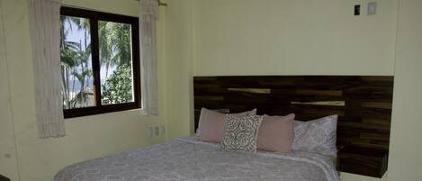 A bedroom with a large bed, a window with curtains, and a view of palm trees.