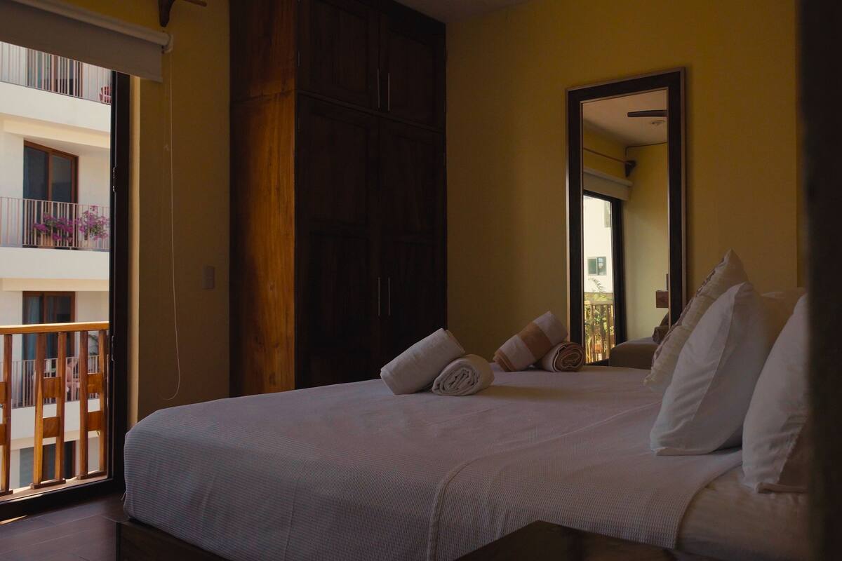 A hotel room with a bed, pillows, a large mirror, and a balcony with a view of a building.