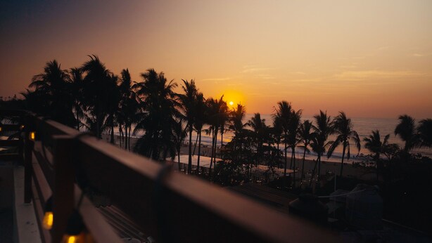 Tramonto visto da un balcone con vista su una spiaggia, palme e un corrimano.