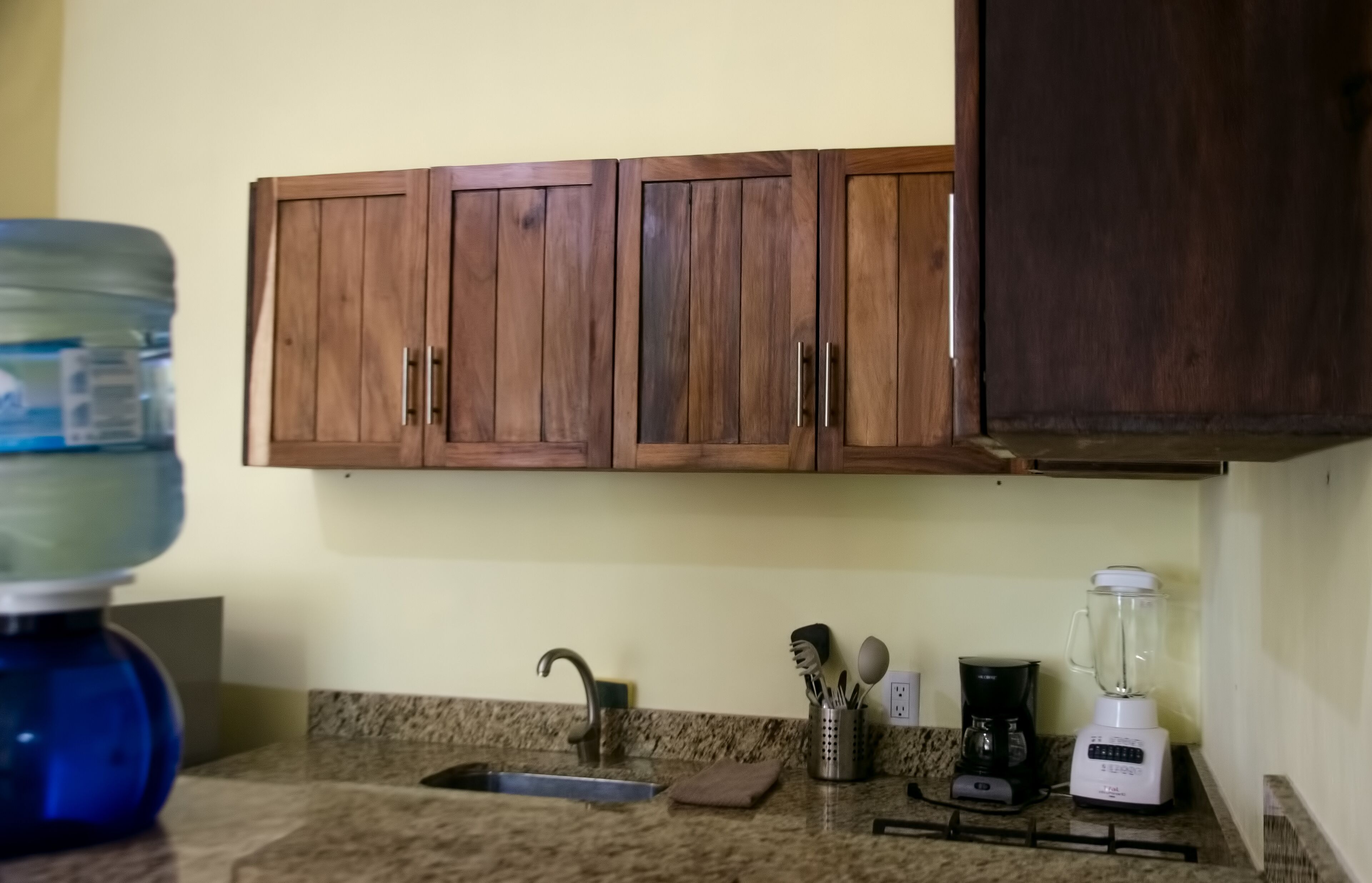 A kitchen with wooden cabinets, a granite countertop, a sink, a coffee maker, a blender, and a water dispenser.