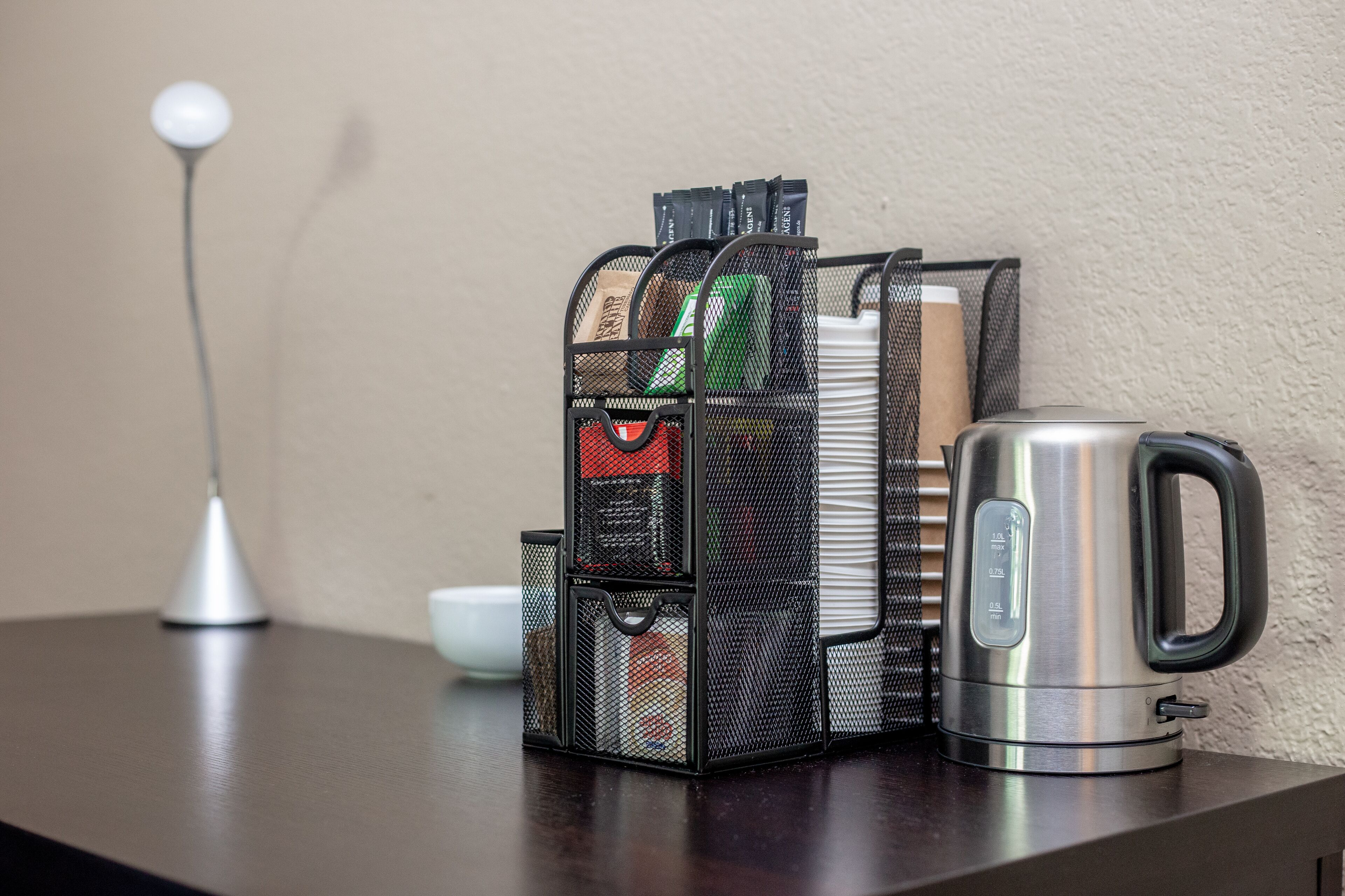 A metal mesh organizer with various items on a dark wooden surface, next to a stainless steel kettle.