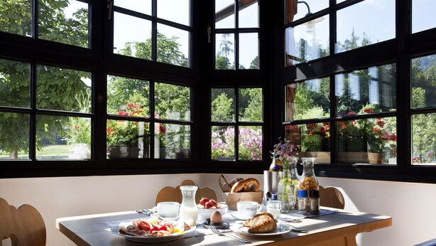 A breakfast table with various food items and drinks, set against a backdrop of large windows with a garden view. - Raitner Wirt (Schleching)