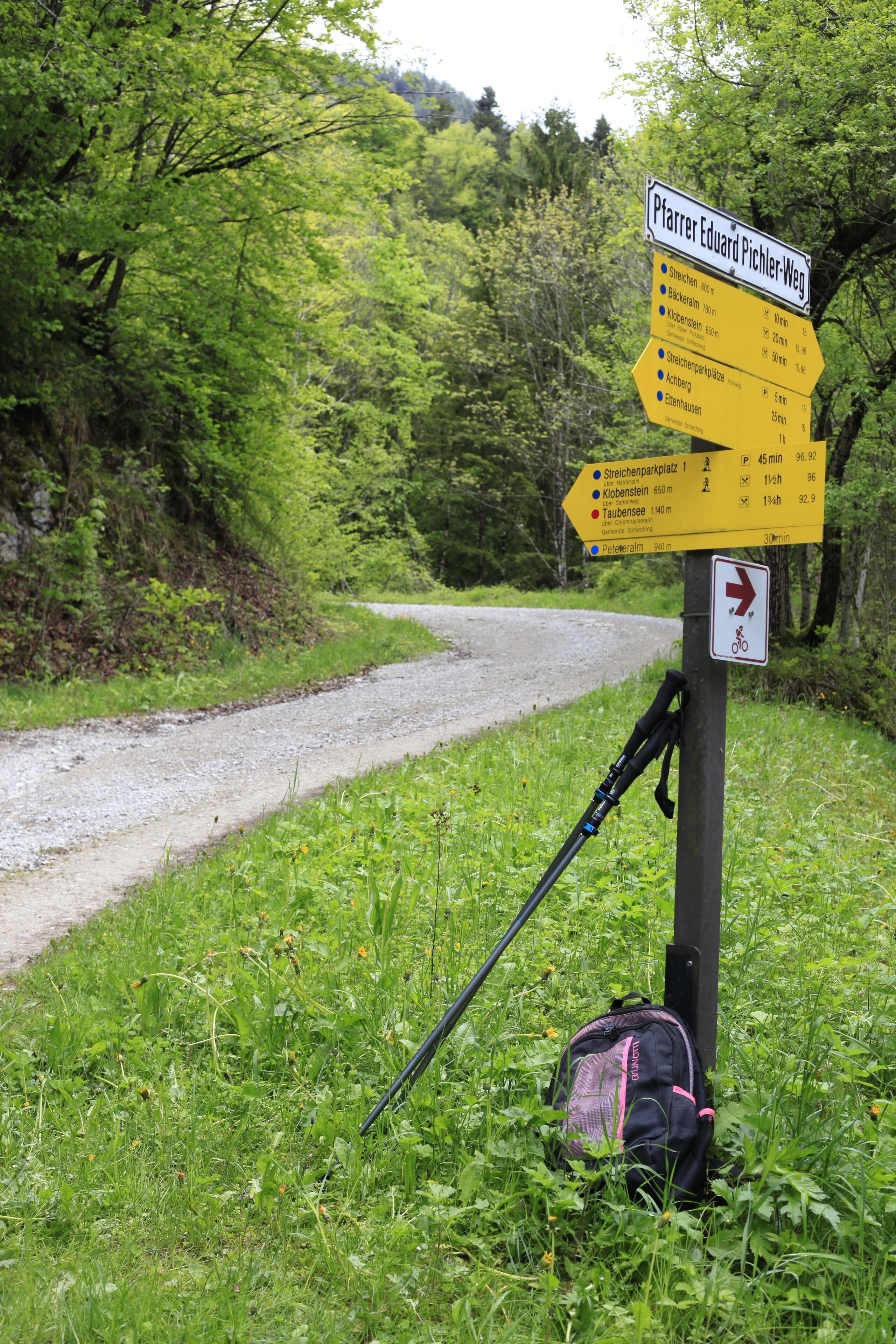 A directional signpost with multiple arrows pointing towards various destinations, a backpack leaning against the post, and a gravel path in a wooded area.