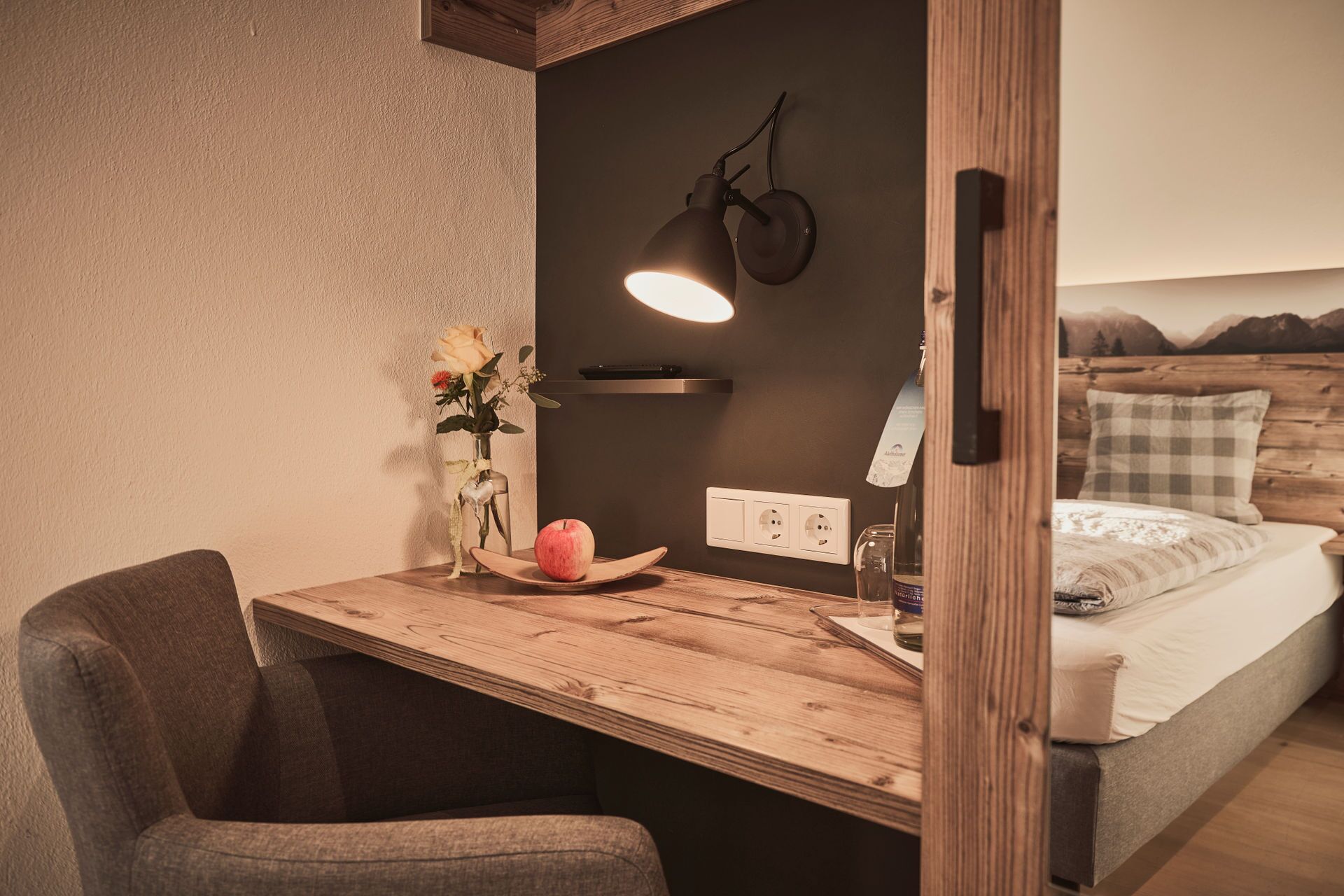 Une chambre confortable avec un bureau en bois, une chaise, une table de chevet avec une lampe, un vase de fleurs et un lit donnant sur une vue sur les montagnes.