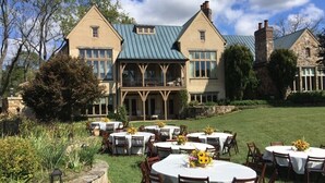 A large building with multiple round tables and chairs set up outdoors, surrounded by greenery and stone walls.